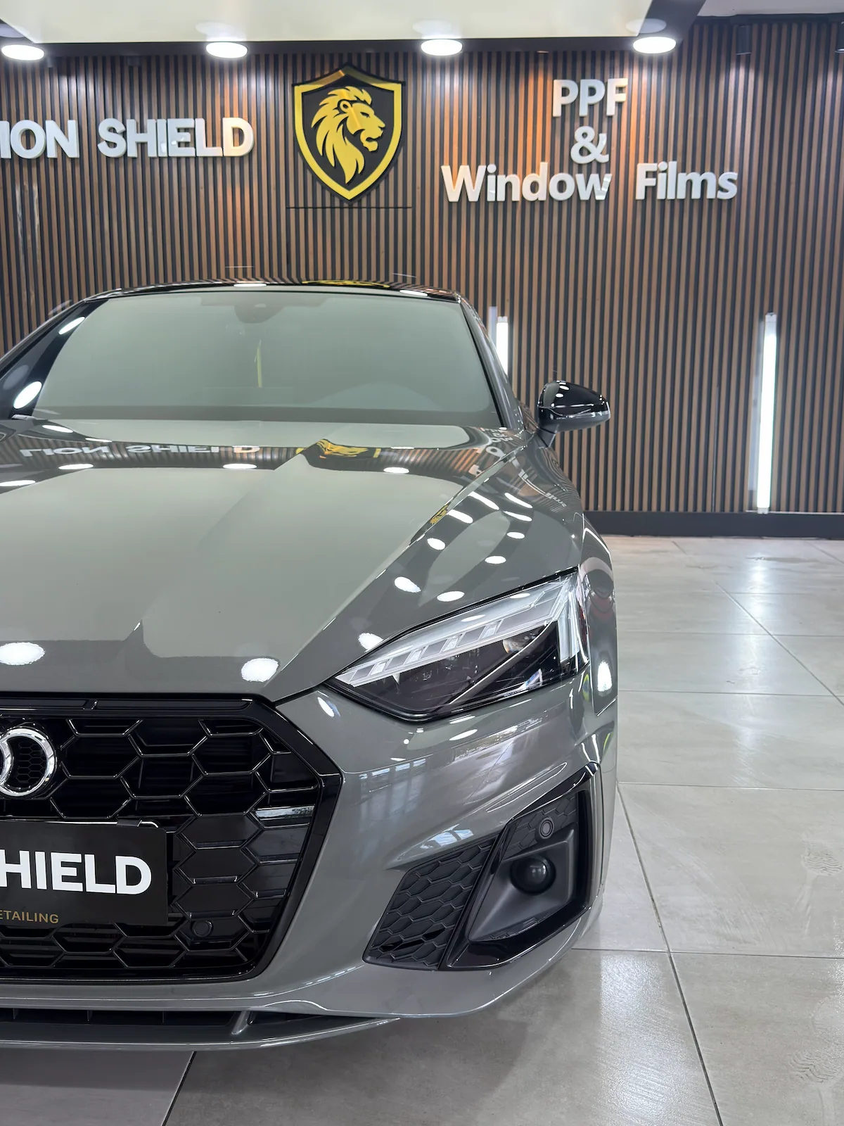 Gri Audi A5’in farı ve kaputu, arkada Lion Shield logolu duvar